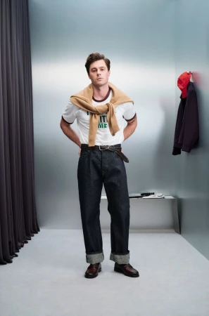 Model in cuffed wide-leg dark denim jeans and a white graphic tee with a camel knit sweater tied around his shoulders in a studio room.