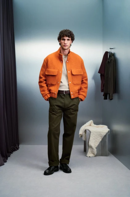 A fair-skinned man poses in an orange jacket and olive pants against a muted blue background with neutral-toned clothes hanging nearby.