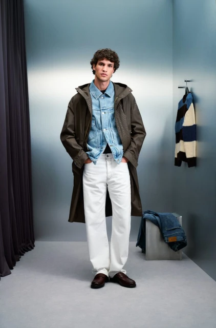 Man in a denim jacket and long olive coat, standing in a studio with white pants and brown shoes, for a fashion editorial shot.