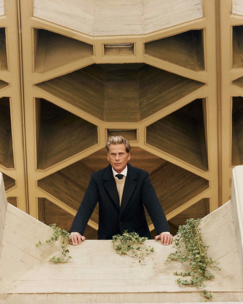 Man in suit standing on balcony with ivy, posing against a geometric wall background, wearing a dark jacket and light sweater.