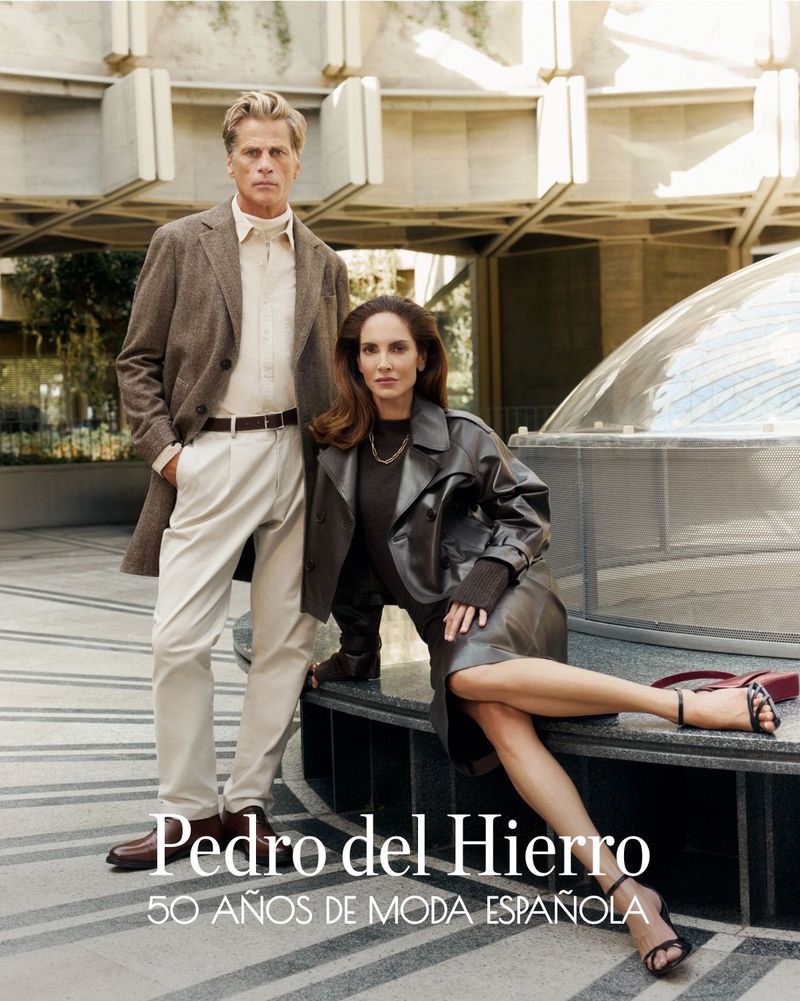 Fashionable couple poses outdoors in front of modern architecture. Man wears brown jacket and white pants, woman in leather coat and dress.
