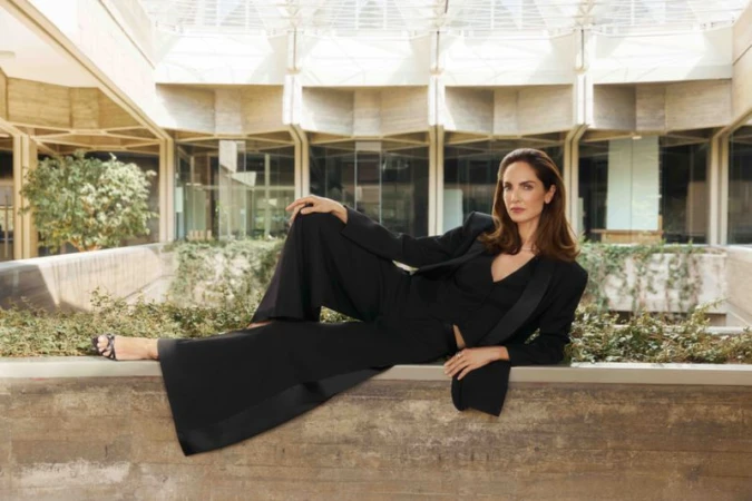 Adult woman in black business suit poses on concrete ledge against a modern building with trees and plants in the background.
