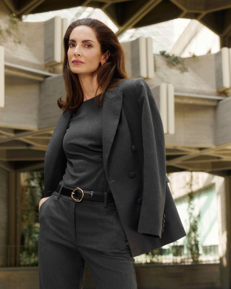 Elegant woman in a grey pantsuit posing against a modern architectural background with natural lighting highlighting her features.