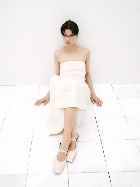 A woman in a light-colored dress and shoes sits on a white tiled floor against a white wall in a studio setting, looking contemplative.