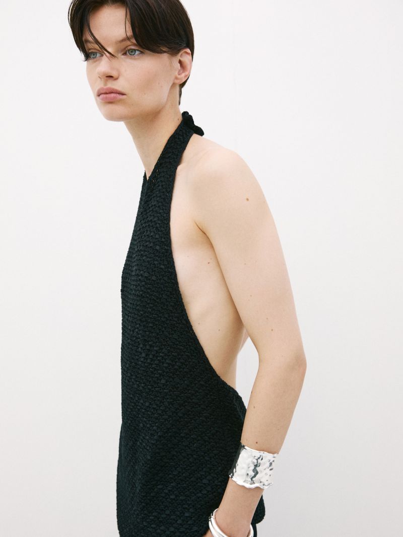 An adult woman poses in a black, textured halter top against a bright white background, also wearing a silver bracelet on her left wrist.
