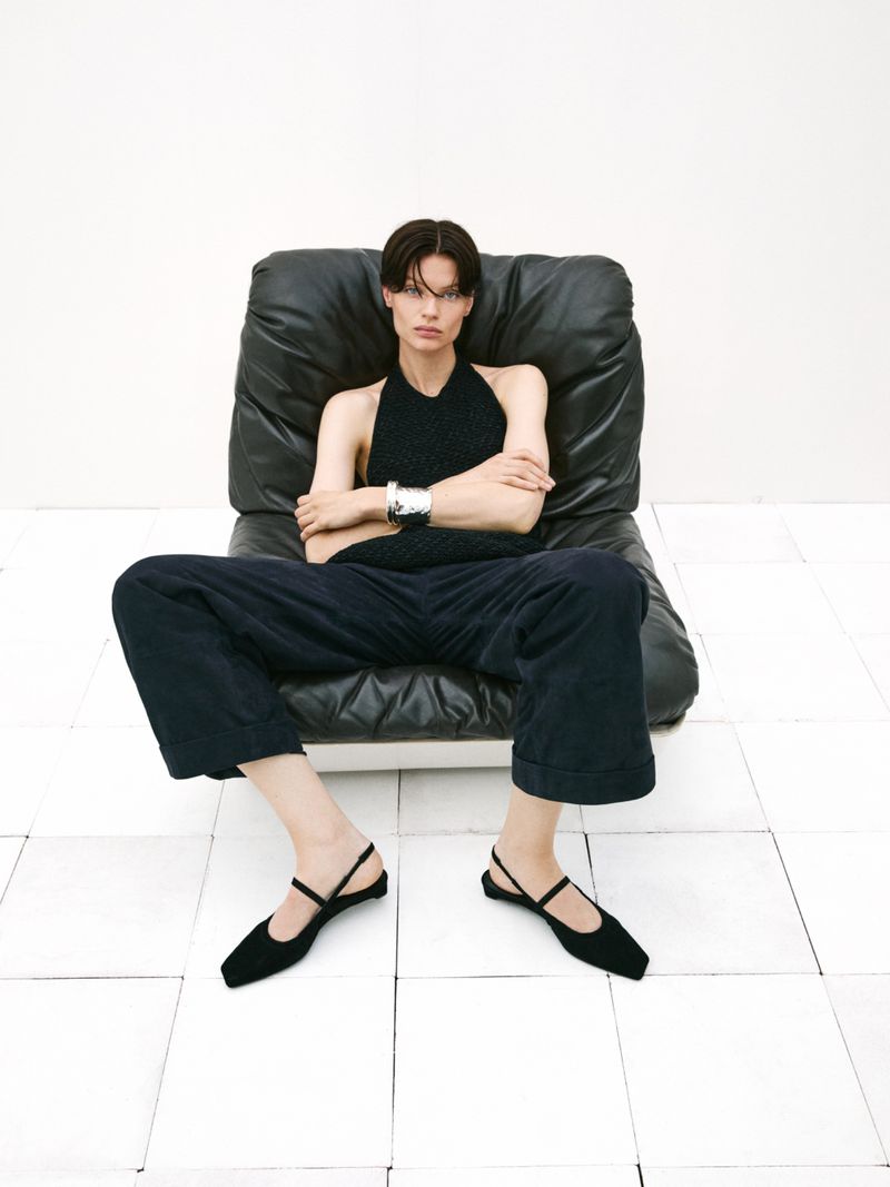 A woman with short dark hair sits in a black leather chair in a minimalist style, wearing a black top, wide-leg trousers, and flats.