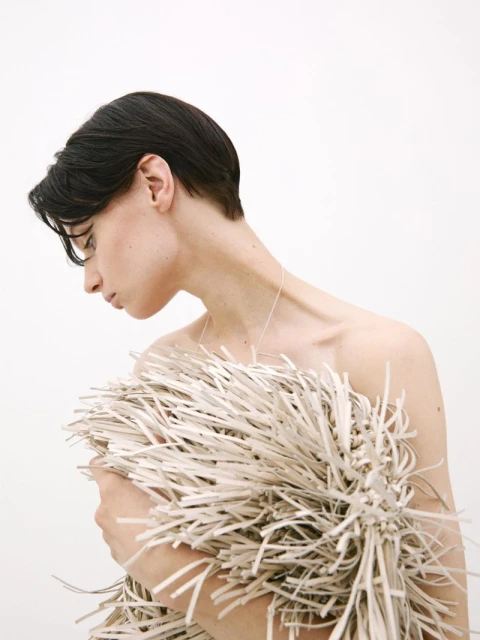 Striking studio portrait of a bare-shouldered young adult, hair cropped short, gazing downward, and wrapped in a cocoon of bundled straw.