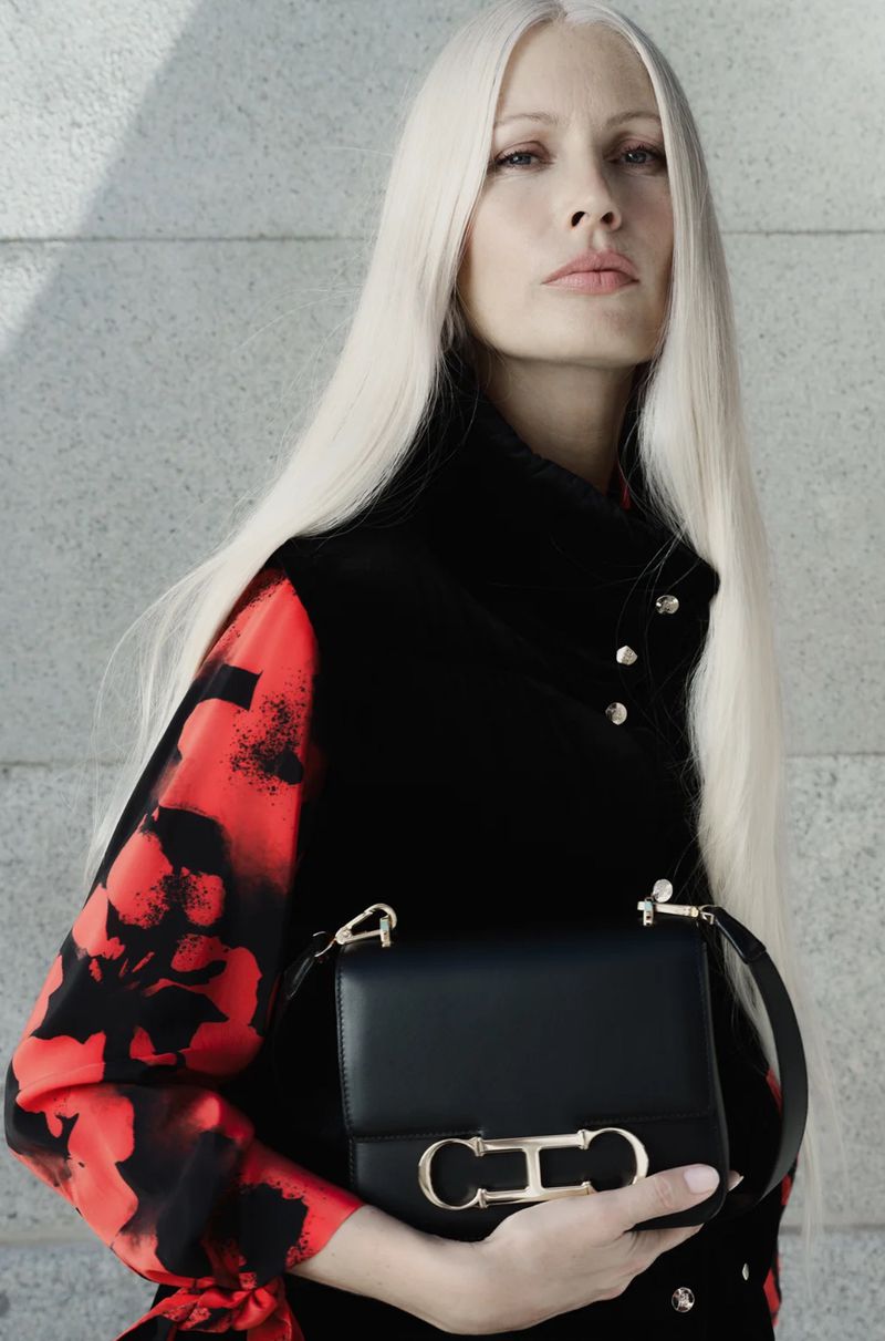 Portrait of a woman with platinum blonde hair holding a black leather purse with a gold clasp against a textured grey wall.