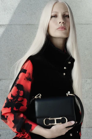 Model with long white hair holding a black leather handbag wearing a red floral blouse and black vest against a stone wall