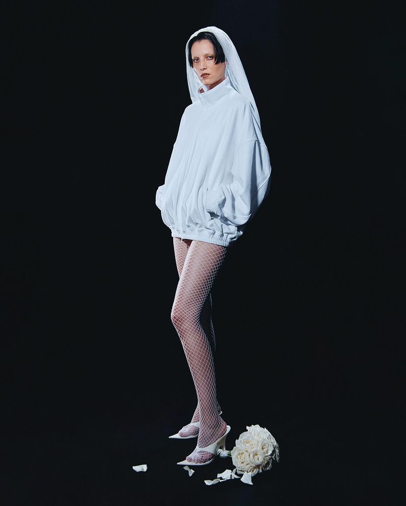 A woman in a white hooded sweatshirt stands in a dark studio with a bouquet of white roses on the floor next to her in a full shot.