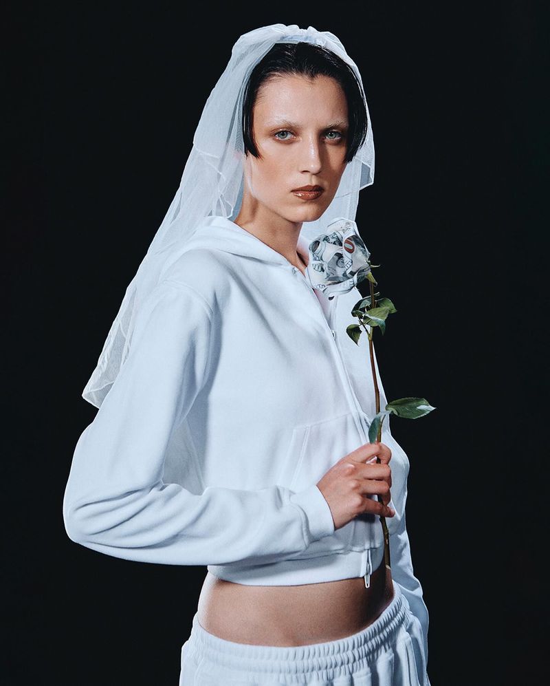 An adult female poses indoors wearing a white hoodie, white pants, and a white bridal veil, holding a silver rose against a dark backdrop.