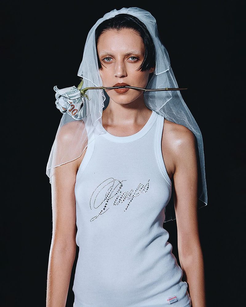 Woman with a rose made of hundred-dollar bills in her mouth, wearing a bridal veil and tank top in a studio setting against a black backdrop.