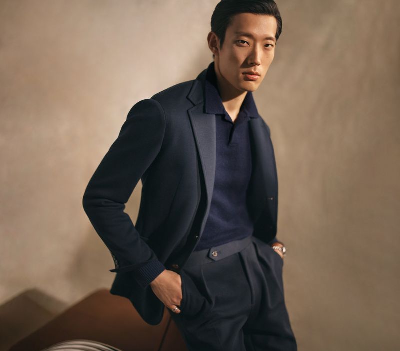 Asian man in navy polo shirt and blazer, posing indoors in elegant suit pants. Fashion portrait with neutral background and soft light.