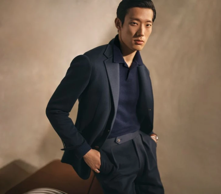 Asian man in navy polo shirt and blazer, posing indoors in elegant suit pants. Fashion portrait with neutral background and soft light.