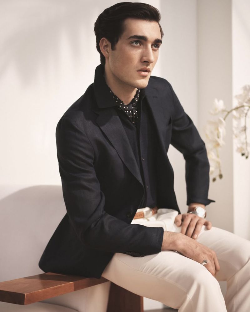 A stylish man sits indoors wearing a black suit jacket, patterned shirt, white pants, and a watch, with decorative flowers in the background.