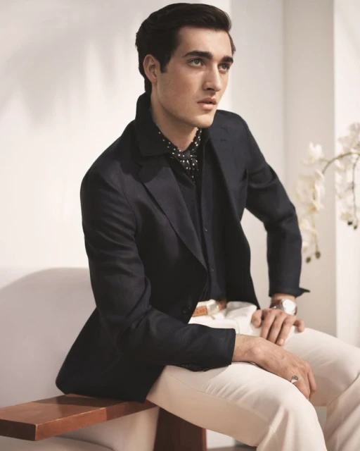 A stylish man sits indoors wearing a black suit jacket, patterned shirt, white pants, and a watch, with decorative flowers in the background.
