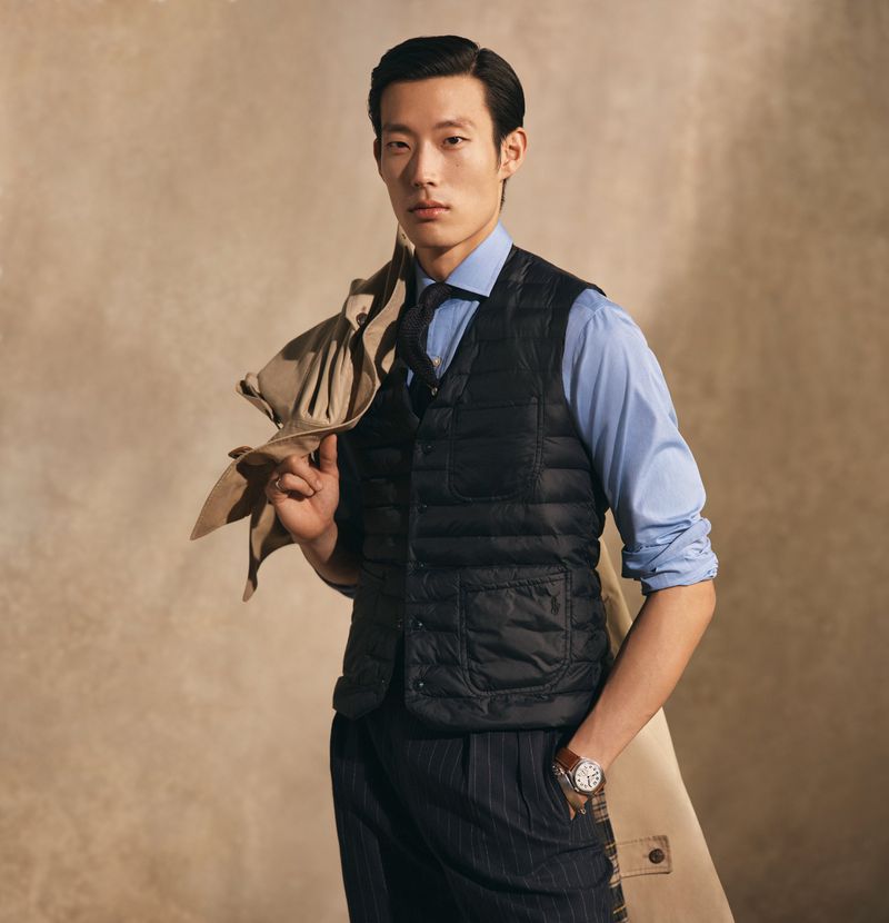 An Asian man is posing indoors. He is wearing a blue button-down shirt, dark vest, and striped trousers. A tan coat is held over his shoulder.