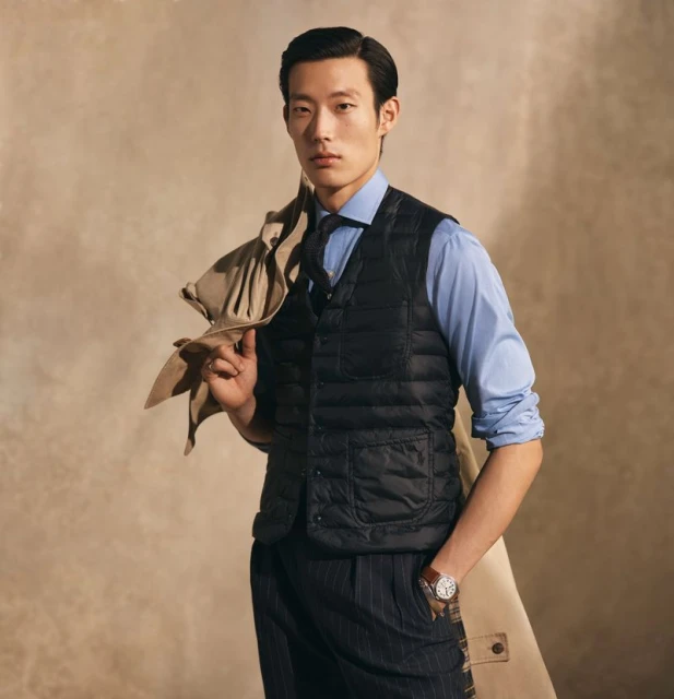 An Asian man is posing indoors. He is wearing a blue button-down shirt, dark vest, and striped trousers. A tan coat is held over his shoulder.