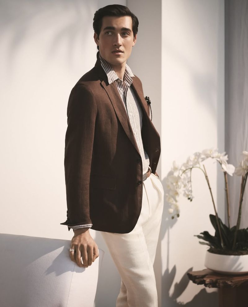 Stylish man in a brown blazer and white pants poses indoors with orchids and a modern chair, exuding fashion and sophistication.