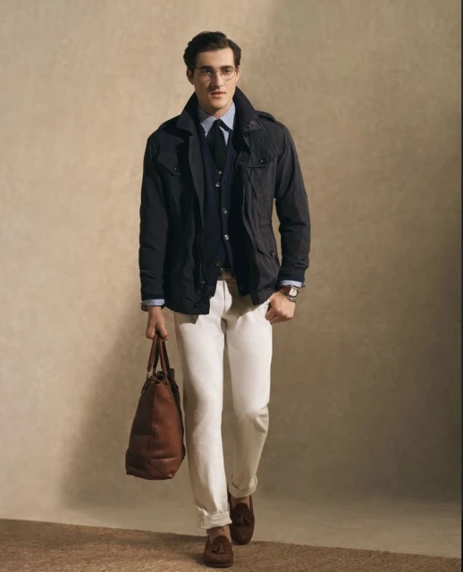 Stylish man in jacket and white pants walks forward carrying a leather tote bag, wearing glasses, against a neutral studio backdrop.