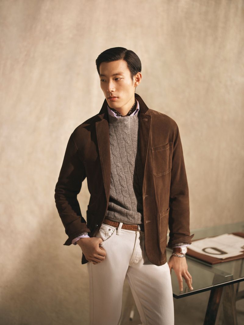 An Asian man in a brown suede jacket, gray cable knit sweater, and light denim jeans poses in an indoor studio setting.