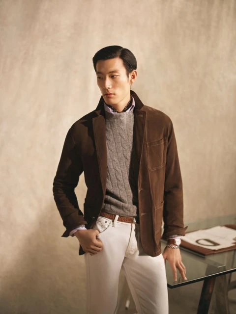 An Asian man in a brown suede jacket, gray cable knit sweater, and light denim jeans poses in an indoor studio setting.