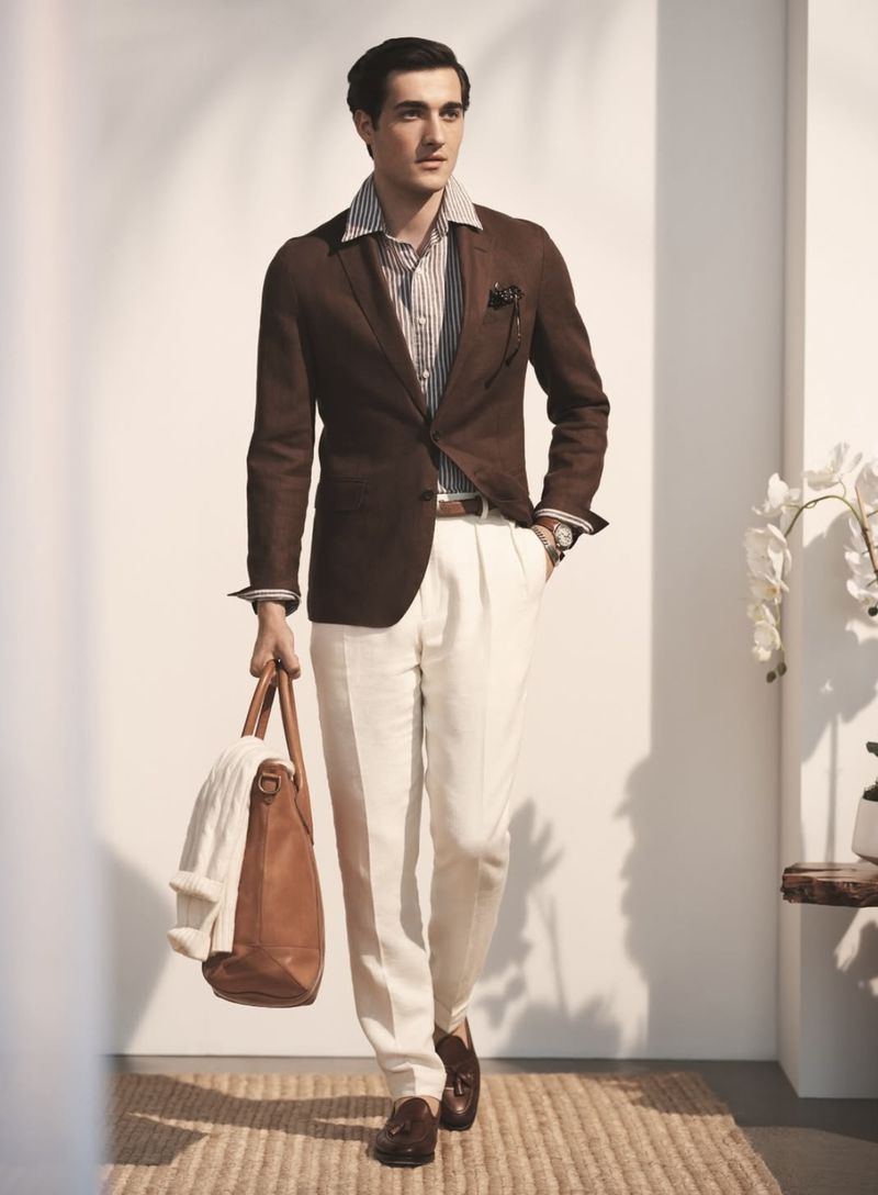 A handsome adult male stands in a light-filled space, wearing a brown linen suit with an open shirt, carrying a leather bag and sweater.