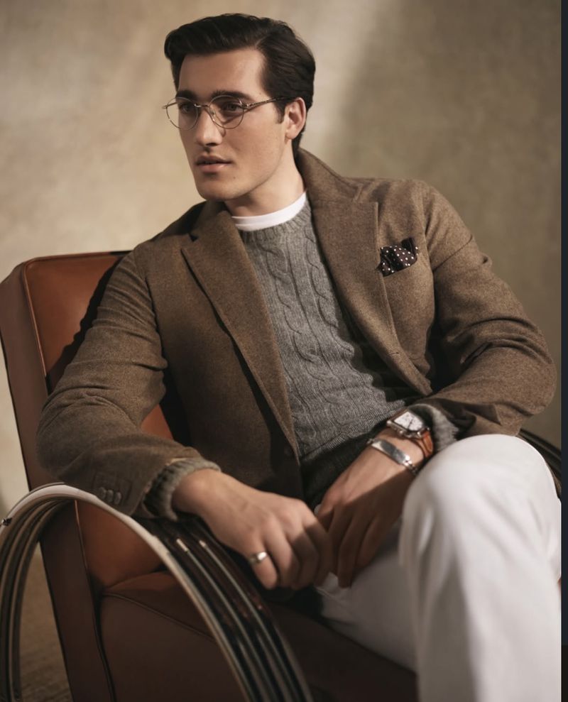 A man wearing a brown blazer and glasses sits in a chair, exuding sophistication in an indoor setting with soft, neutral lighting.