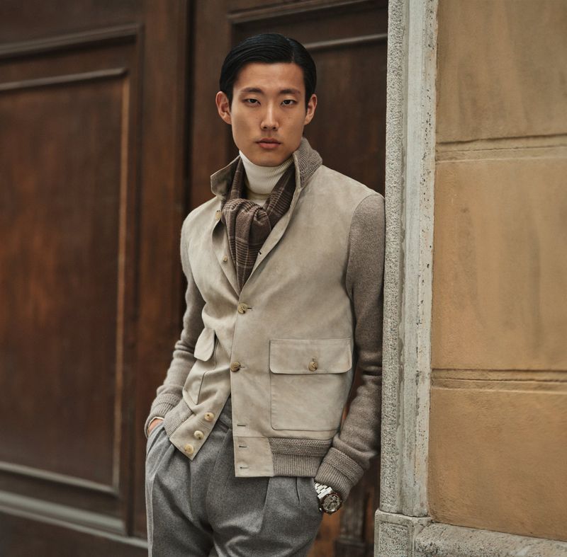 Asian man poses wearing tan jacket and grey slacks outdoors in front of wooden doors. Fall fashion editorial shot with natural light.
