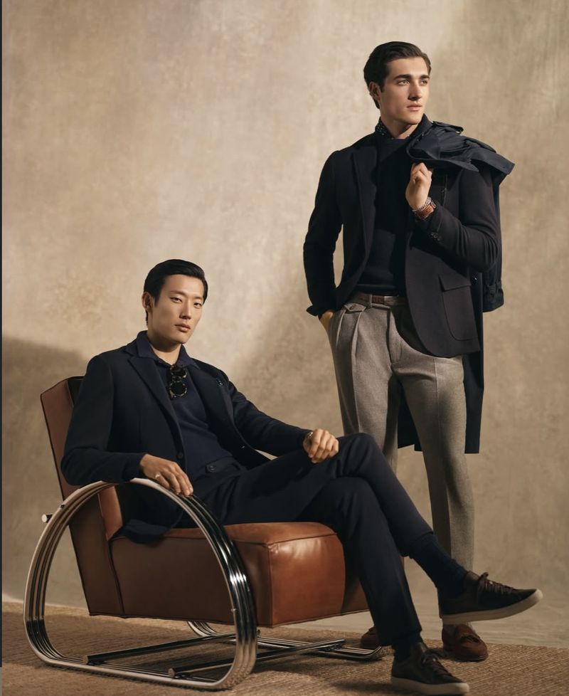 Two men in stylish dark blazers and dress pants pose indoors against a neutral backdrop. One sits in a leather chair.