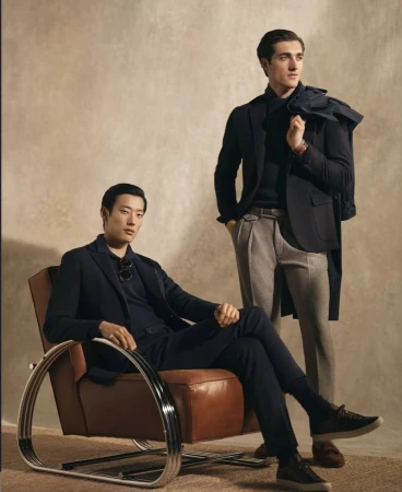 Two men in stylish dark blazers and dress pants pose indoors against a neutral backdrop. One sits in a leather chair.