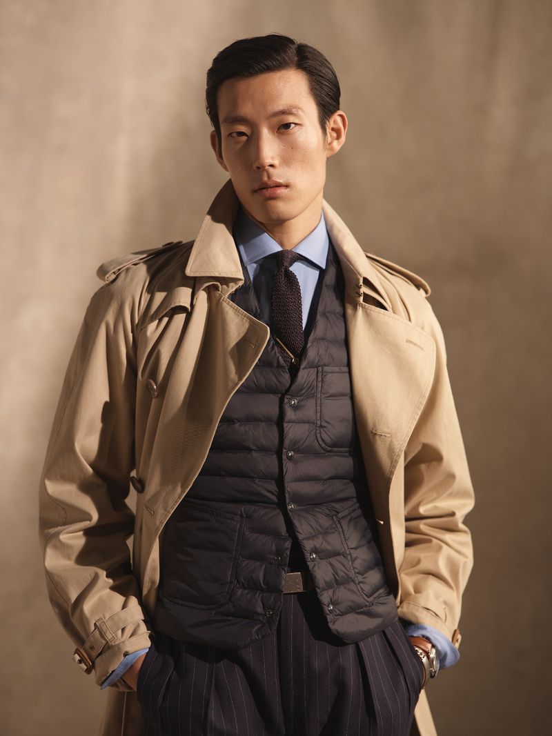 Portrait of a handsome Asian man in a trench coat and puffer vest, posing indoors with a neutral backdrop, showcasing a fashionable style.