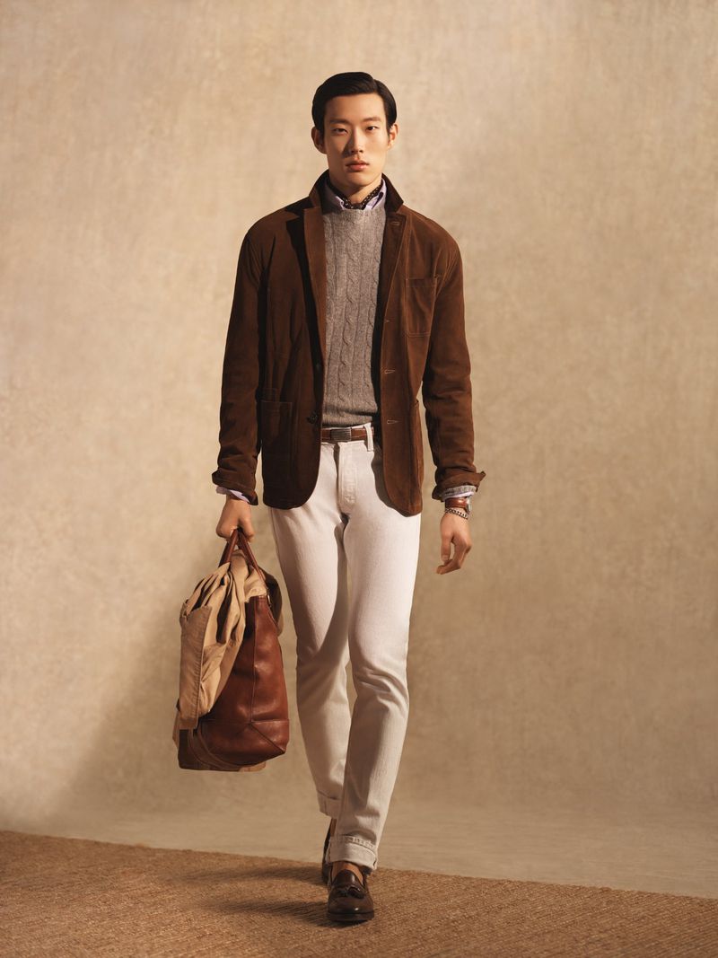 Model Juhyung Kang walking with a brown leather duffle bag wearing a brown jacket, grey cable knit sweater, and white trousers.