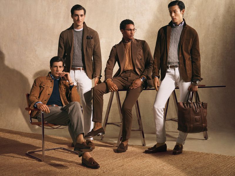 Four adult men in casual-yet-stylish attire pose indoors; three stand and one sits in a chair, with neutral background. Fashion shoot.