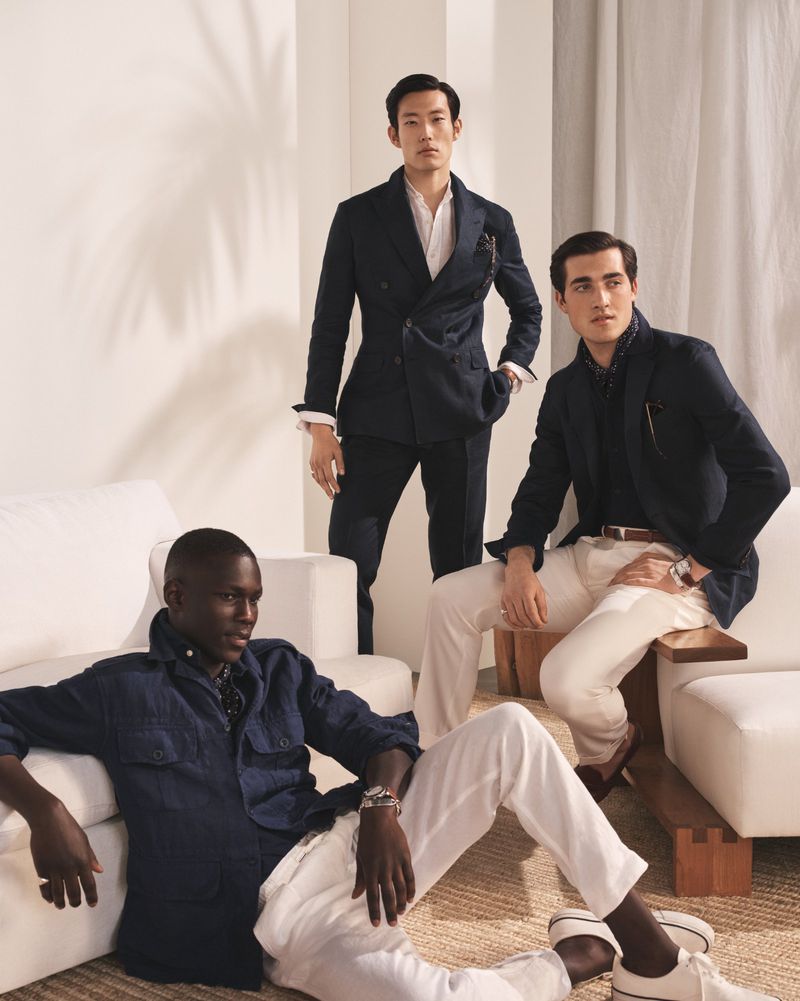 Three male fashion models in blazers and pants are posed indoors against a white backdrop, sitting and standing on a woven rug.