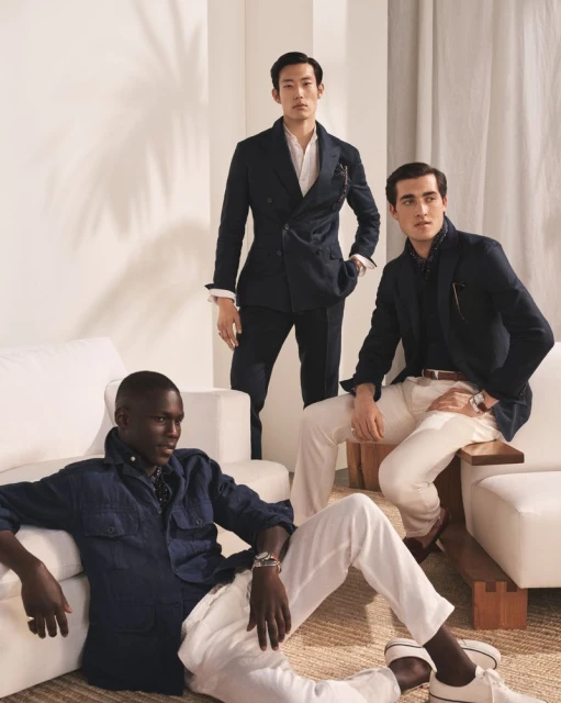 Three male fashion models in blazers and pants are posed indoors against a white backdrop, sitting and standing on a woven rug.