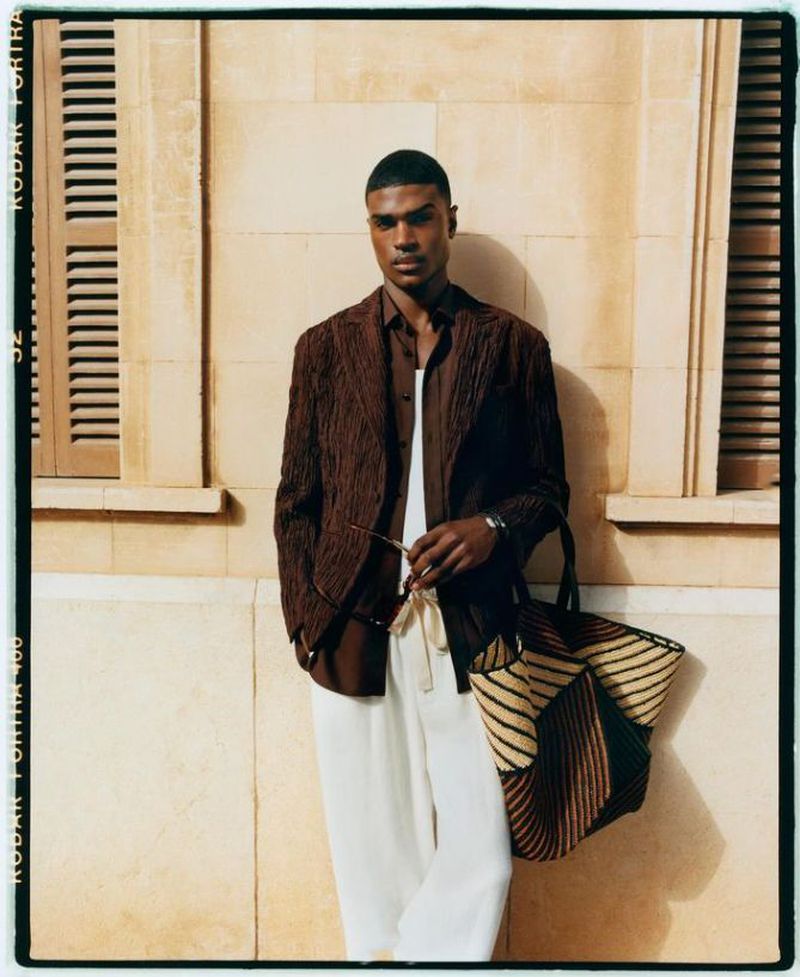 Portrait of a stylish adult Black man in a brown jacket and white pants, holding sunglasses and a woven tote bag indoors.