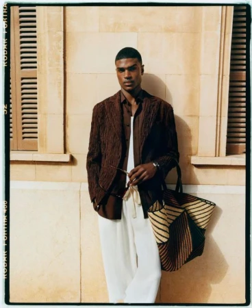 Portrait of a stylish adult Black man in a brown jacket and white pants, holding sunglasses and a woven tote bag indoors.