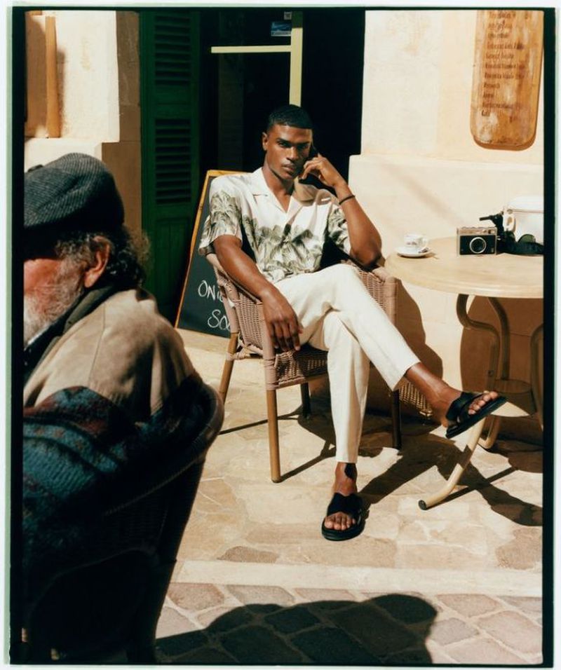 A stylish dark-skinned man in a patterned shirt and white pants sits outside at a cafe table with a film camera and coffee.