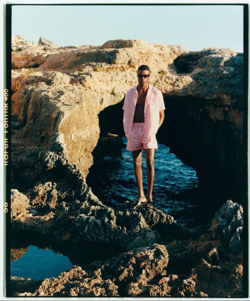A man standing on rocky shore wearing a pink striped shirt and shorts near the ocean.
