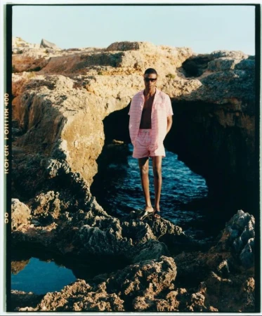 A man standing on rocky shore wearing a pink striped shirt and shorts near the ocean.