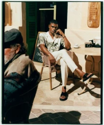 Model Rafael Mayers sitting outside a cafe in a printed short-sleeve shirt, cream trousers, and black slide sandals.