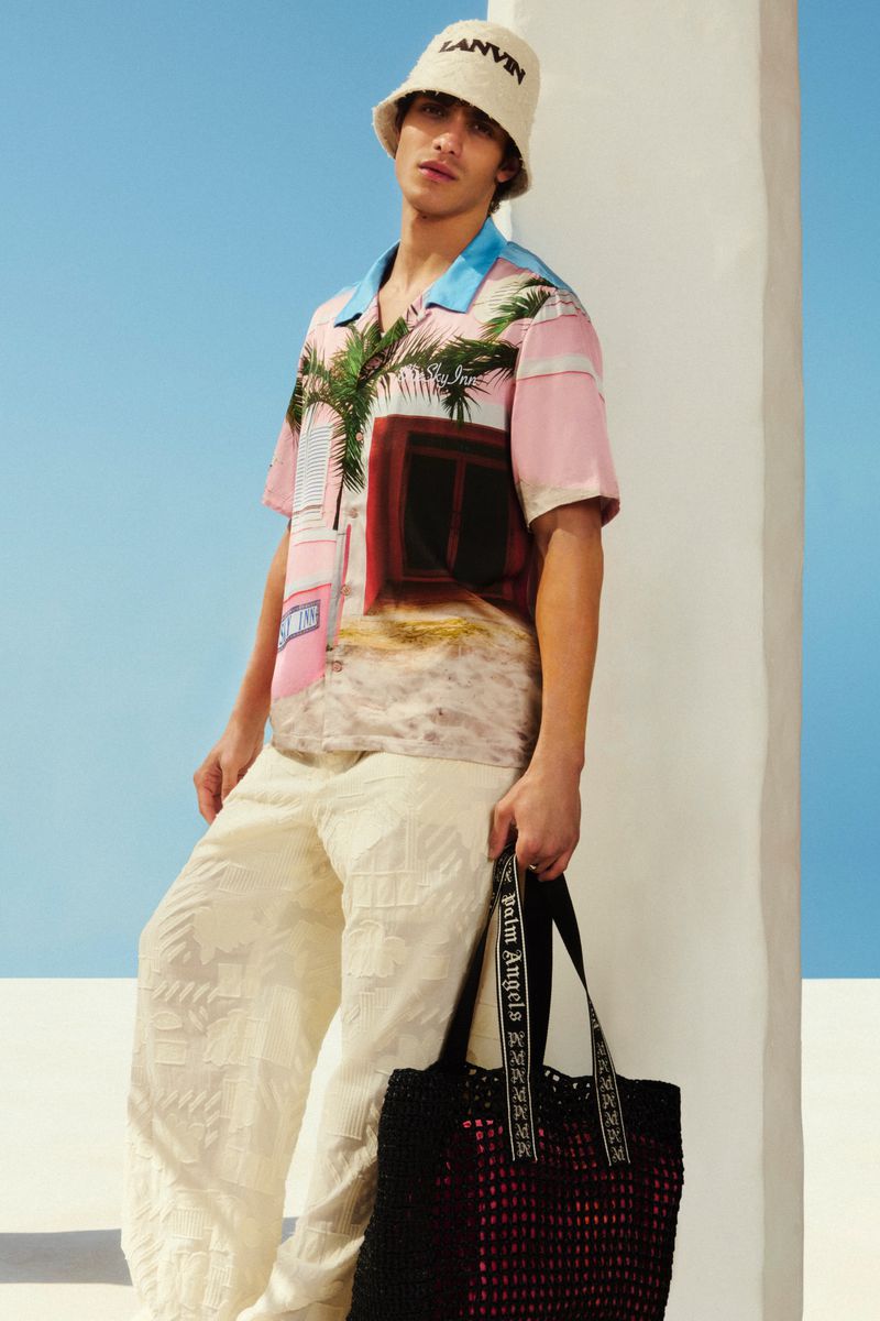 Man in patterned shirt and bucket hat holds black tote bag in outdoor setting with blue sky. Fashionable summer style.