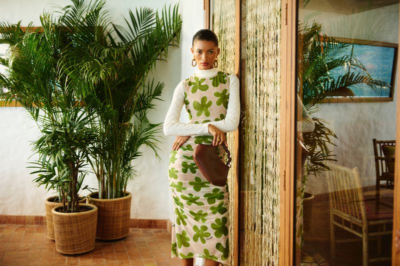 Indoor fashion portrait of an adult woman in a flower-print dress, posing next to a palm tree in a pot, styled with a brown bag.