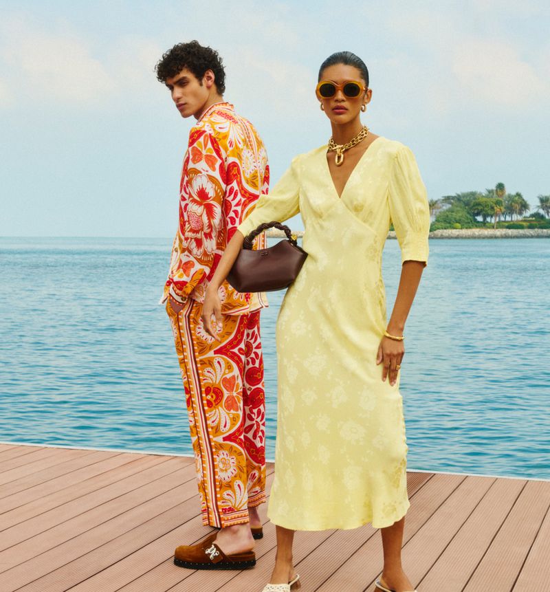 Two people are posing on a wooden dock next to the ocean. The woman wears a yellow dress and the man wears a patterned orange suit.