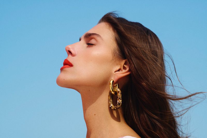 Close-up profile of a woman with red lipstick and gold earrings against a clear blue sky, looking upwards with her eyes closed.