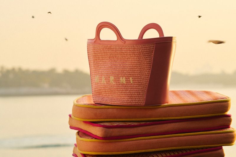 Fashion shot of a handbag on top of cushions with a background of a seashore, and distant land under a sunset-colored sky.