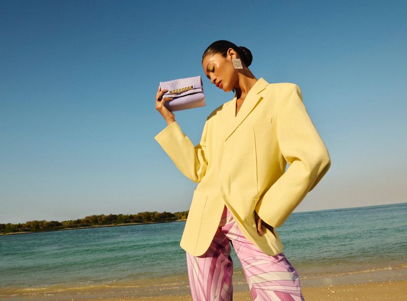 Woman in yellow blazer and patterned pants poses on beach with lilac purse, clear blue sky and ocean in the background.