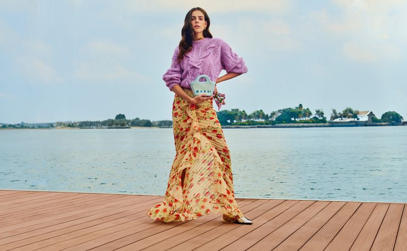 A woman poses on a wooden platform with a floral skirt, lilac sweater, and light blue purse, against a waterfront background and cloudy sky.