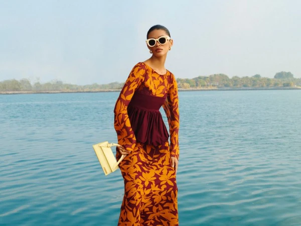 A woman poses in a floral dress and sunglasses, holding a purse, with a blue ocean backdrop under a clear sky. Fashion shot.